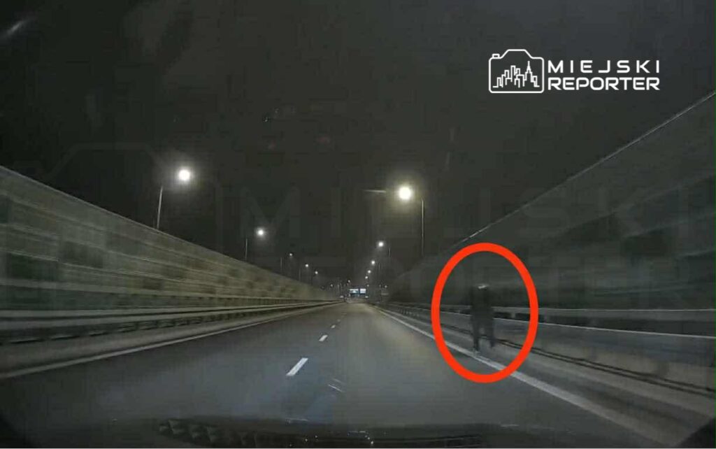 Osoba w ciemnym ubraniu idąca wzdłuż drogi na nocnej trasie, oświetlonej latarniami.