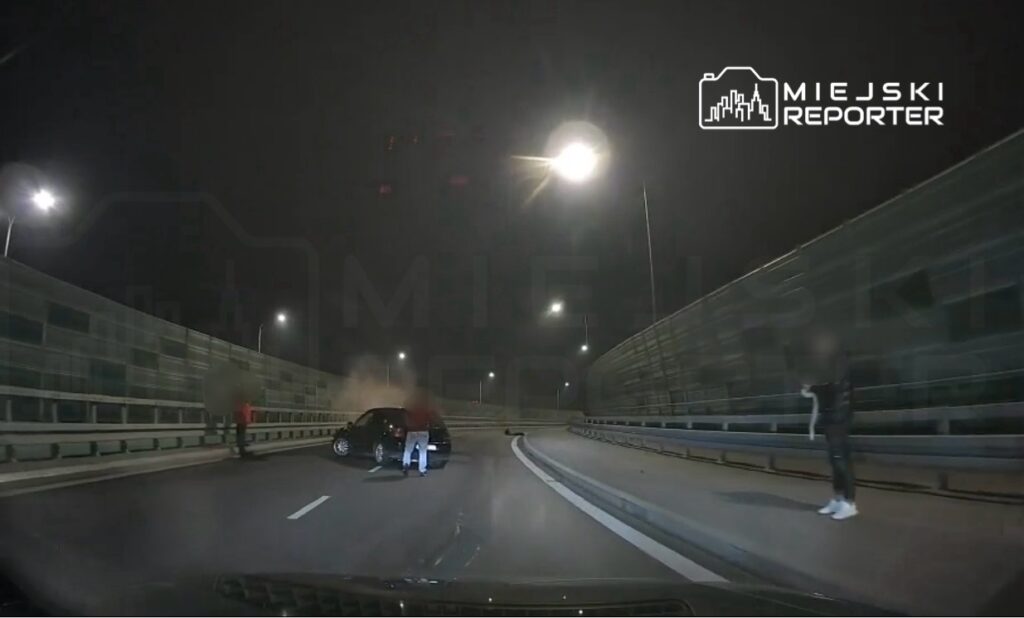 Czarna osobówka stoi na drodze, a obok niej grupa osób w nocy, w tle widoczne latarnie.