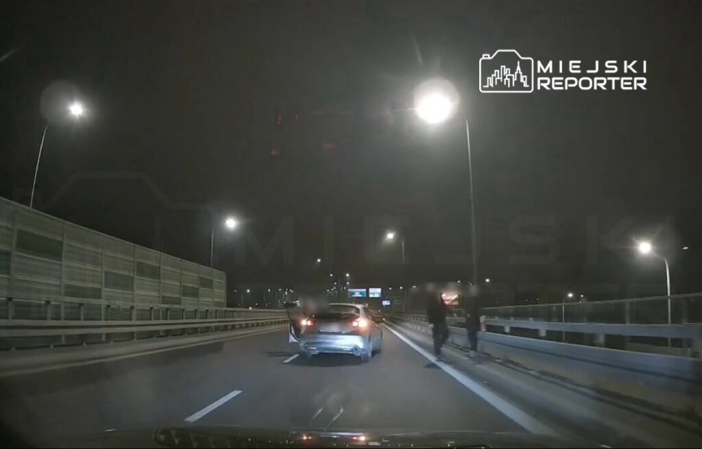 Na drodze ekspresowej nocą, samochód osobowy zatrzymany z otwartymi drzwiami, obok idący pieszy.