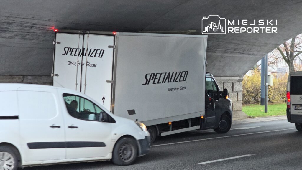 Ciężarówka z logo "SPECIALIZED" przejeżdża pod niskim wiaduktem drogowym, obok białego vana.