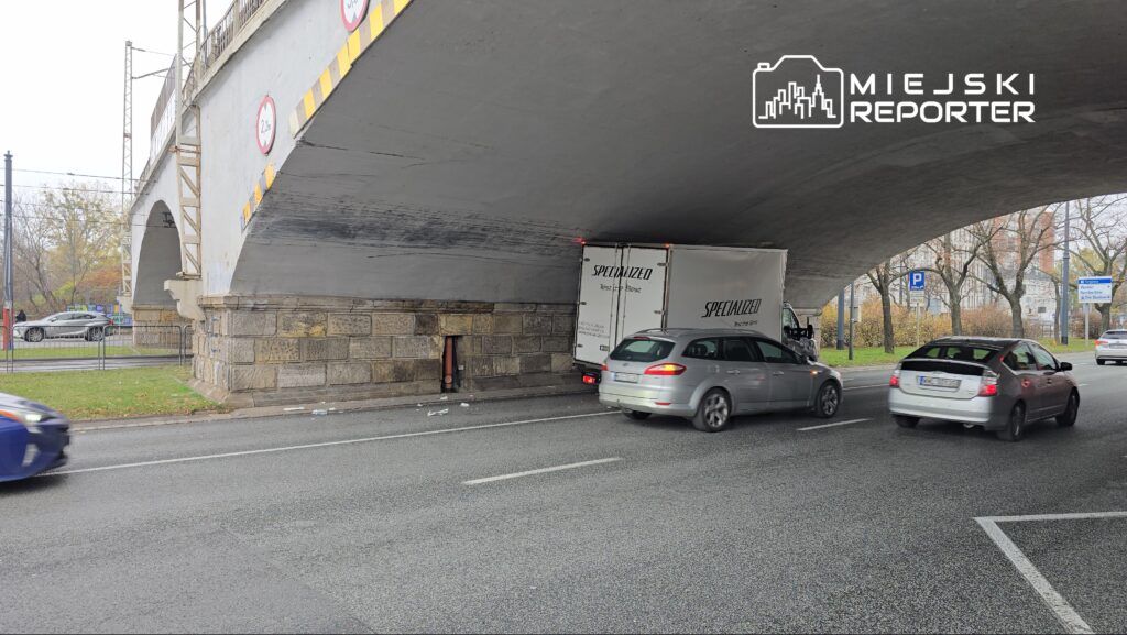 Niski most, pod którym utknął dostawczy samochód, obok poruszają się inne pojazdy na ruchliwej ulicy.