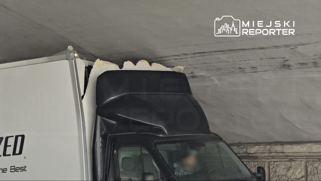 Ciężarówka z uszkodzonym dachem stoi pod niskim stropem, widać zniszczenia na górnej krawędzi pojazdu.