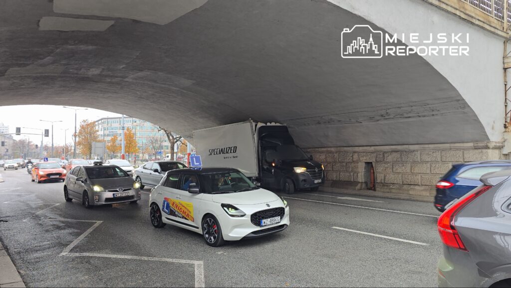 Ruch samochodowy pod wiaduktem, z ciężarówką utknętą w wąskim miejscu, w tle nowoczesne budynki i drzewa.