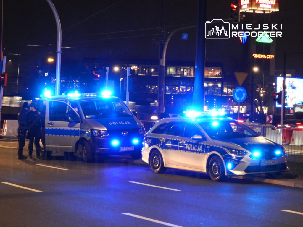 Policjanci rozmawiają przy radiowozie z włączonymi sygnałami świetlnymi na miejskiej ulicy w nocy.
