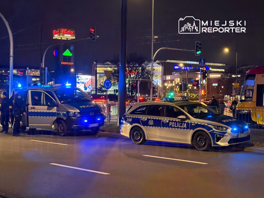 Policjanci w mundurach wychodzą z radiowozu na oświetlonej ulicy, obok widoczne są reklamy i światła miasta.