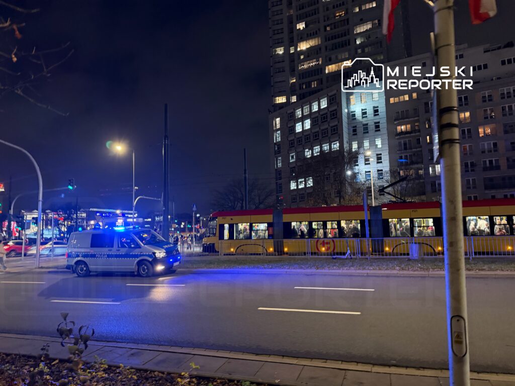 Policyjny samochód z niebieskimi światłami przejeżdża obok tramwaju pełnego pasażerów w nocy.