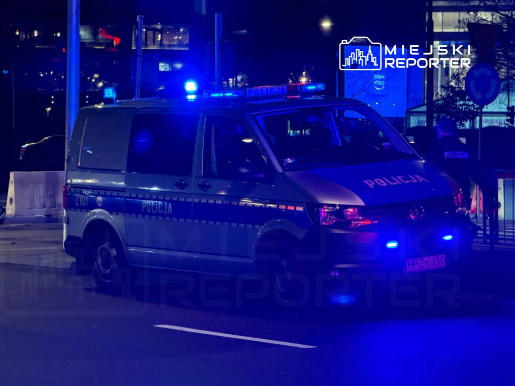 Policyjny samochód z włączonymi sygnałami świetlnymi zatrzymany na ulicy w nocy, w tle widać oświetlone budynki.
