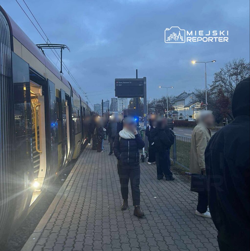 Grupa pasażerów czeka na przystanku tramwajowym, obok nowoczesnego tramwaju, w otoczeniu miejskich budynków.