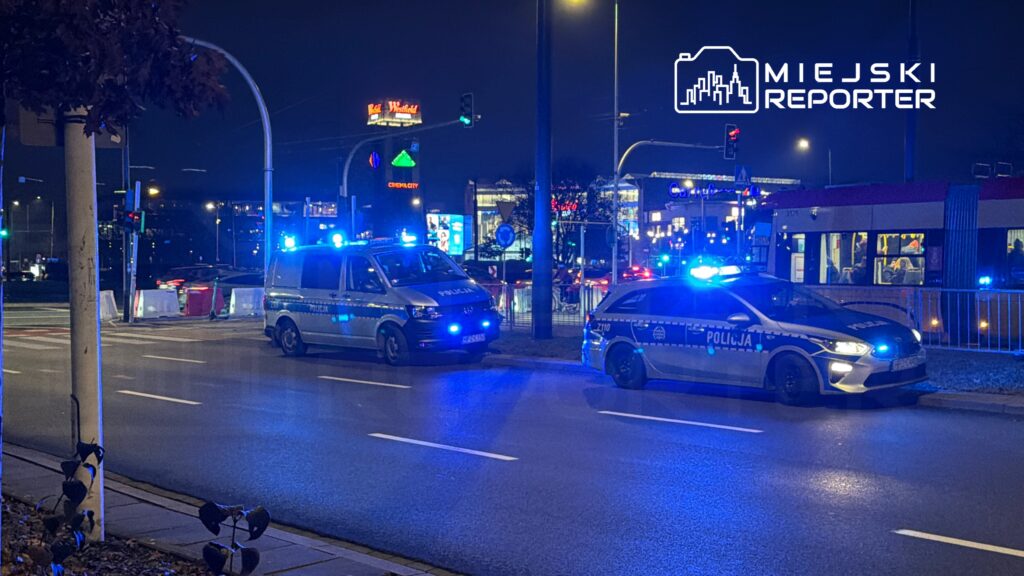 Dwa radiowozy policyjne z włączonymi sygnałami świetlnymi zatrzymane przy ruchliwej ulicy w nocy.