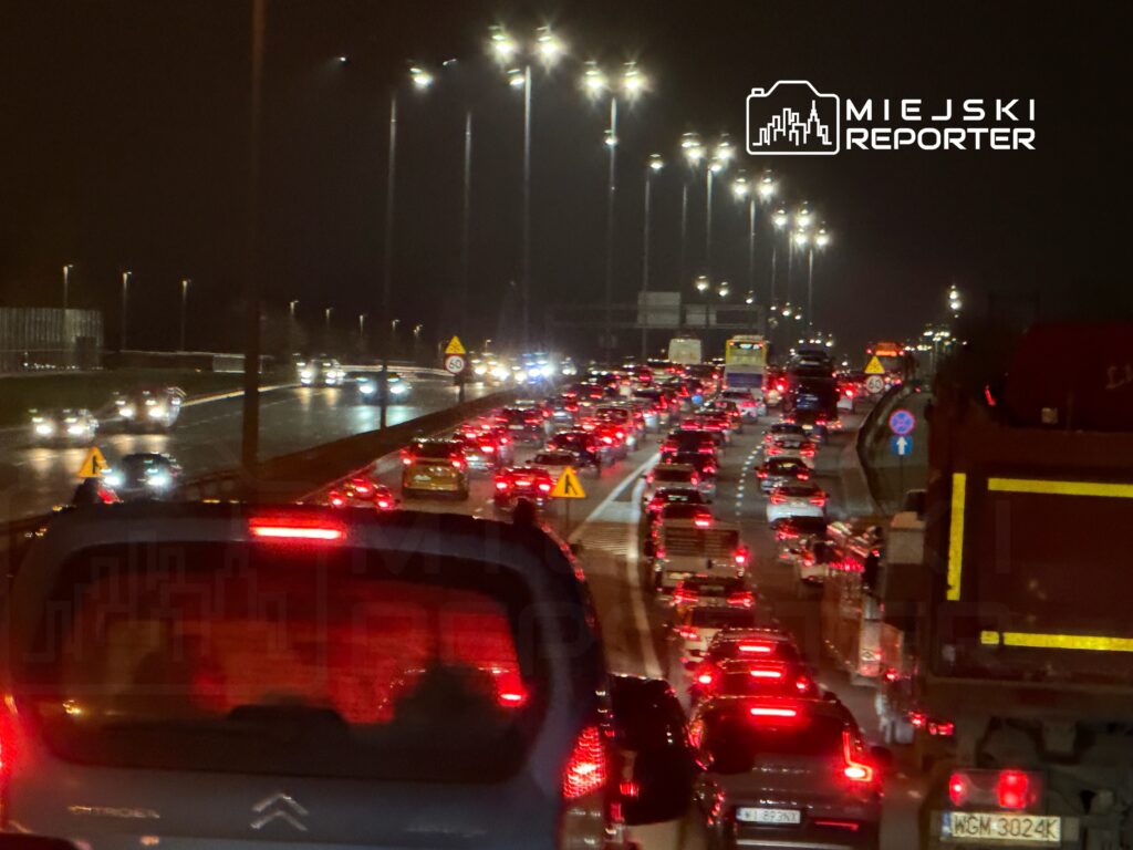 Ruch samochodowy w godzinach wieczornych na oświetlonej drodze, z wieloma pojazdami stojącymi w korku.