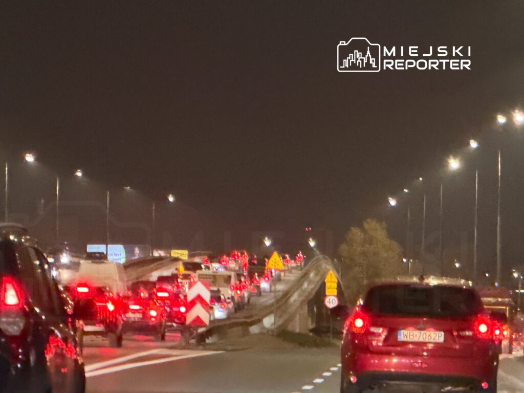 Korkujący ruch samochodowy na drodze ekspresowej w nocy, z widocznymi światełkami pojazdów i znakami drogowymi.