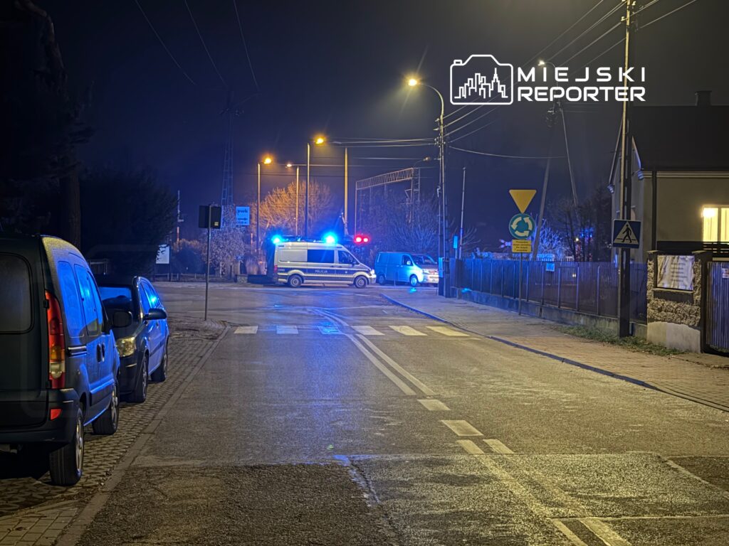 Na ulicy nocą stoi radiowóz policyjny z włączonymi światłami, obok zaparkowane samochody.