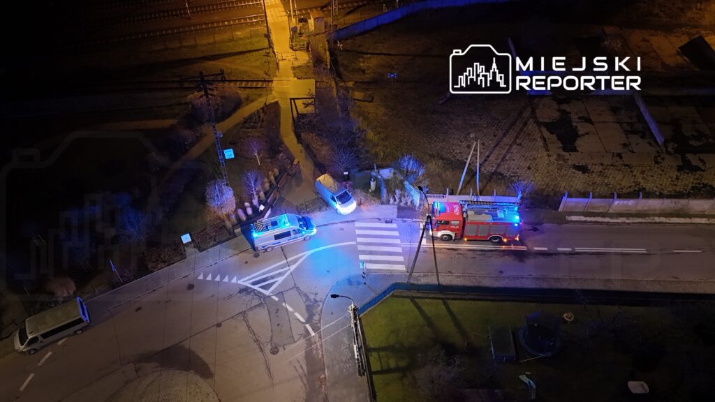 W nocy na skrzyżowaniu ulicy stoją samochody policyjne i wóz strażacki z włączonymi sygnałami świetlnymi.