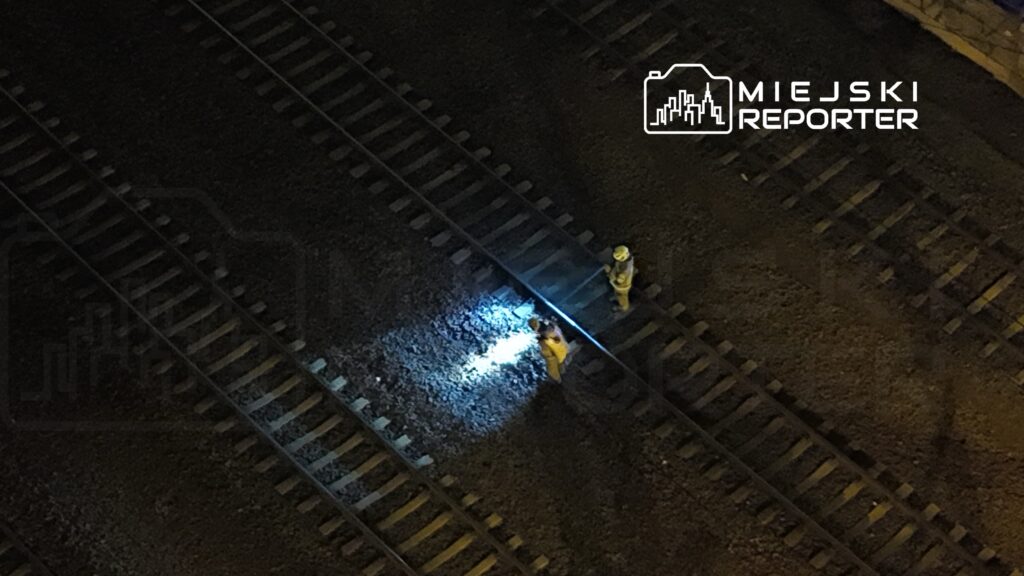 Dwaj pracownicy w żółtych kombinezonach sprawdzają tory kolejowe w nocy, używając latarki.