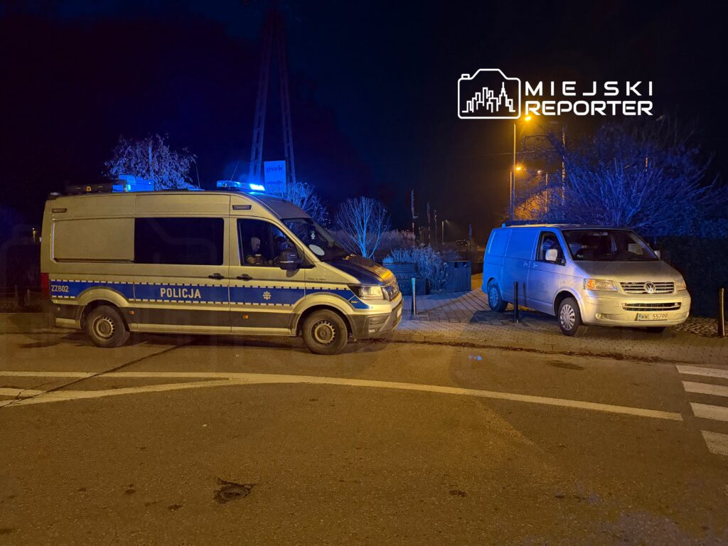 Policyjny furgon z niebieskimi światłami zatrzymany na drodze, obok stoi szary van. Nocna sceneria z oświetleniem ulicznym.