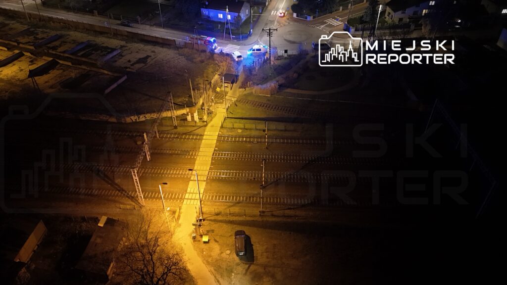 Nieoznakowany radiowóz policyjny zatrzymany przy torach kolejowych w nocy, oświetlony niebieskimi światłami.