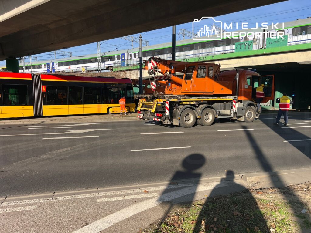 Pomarańczowy dźwig podnosi tramwaj, podczas gdy pracownicy w odblaskowych kamizelkach kierują ruchem na drodze.