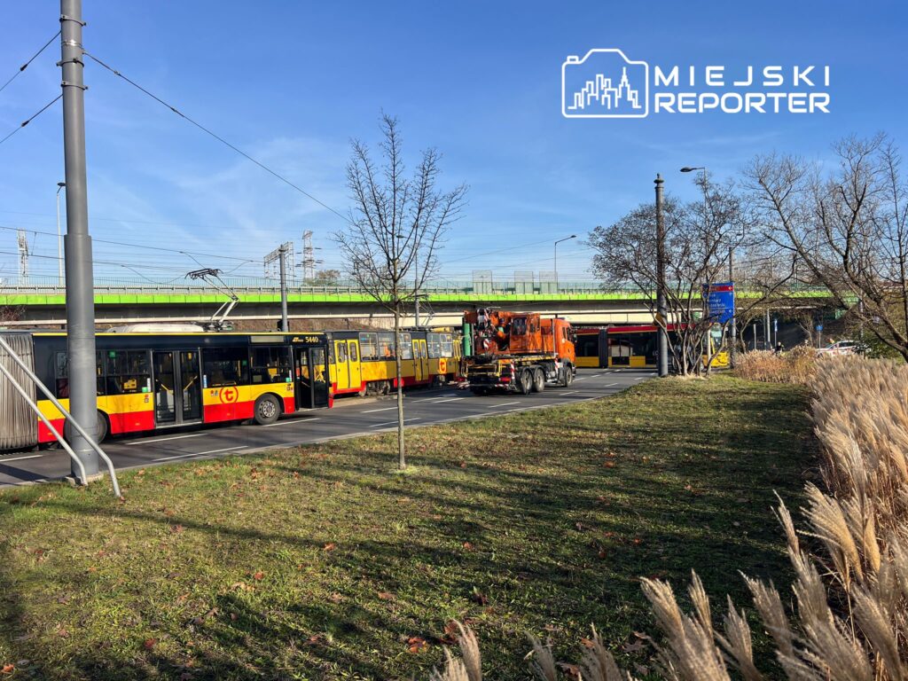 Tramwaje w barwach żółto-czerwonych stoją na przystanku obok drogi, w tle widoczny most i niebieskie niebo.