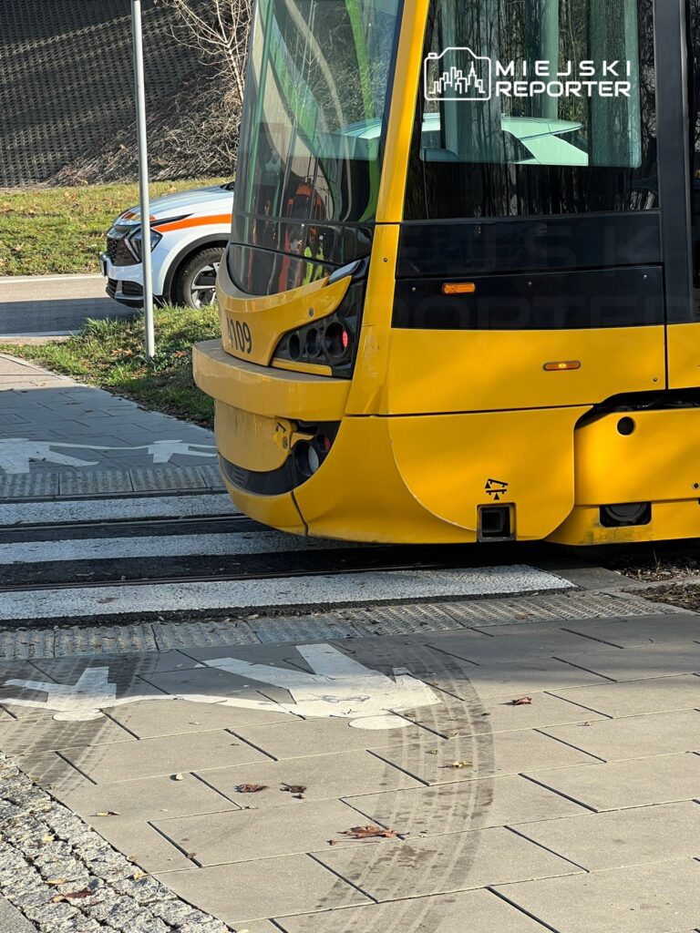 Żółty tramwaj zatrzymany na przystanku w miejskim otoczeniu, obok widoczny samochód służbowy.