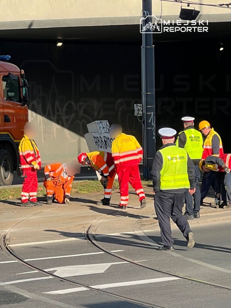 Grupa pracowników w odblaskowych kamizelkach prowadzi prace naprawcze na torowisku tramwajowym w miejskim otoczeniu.