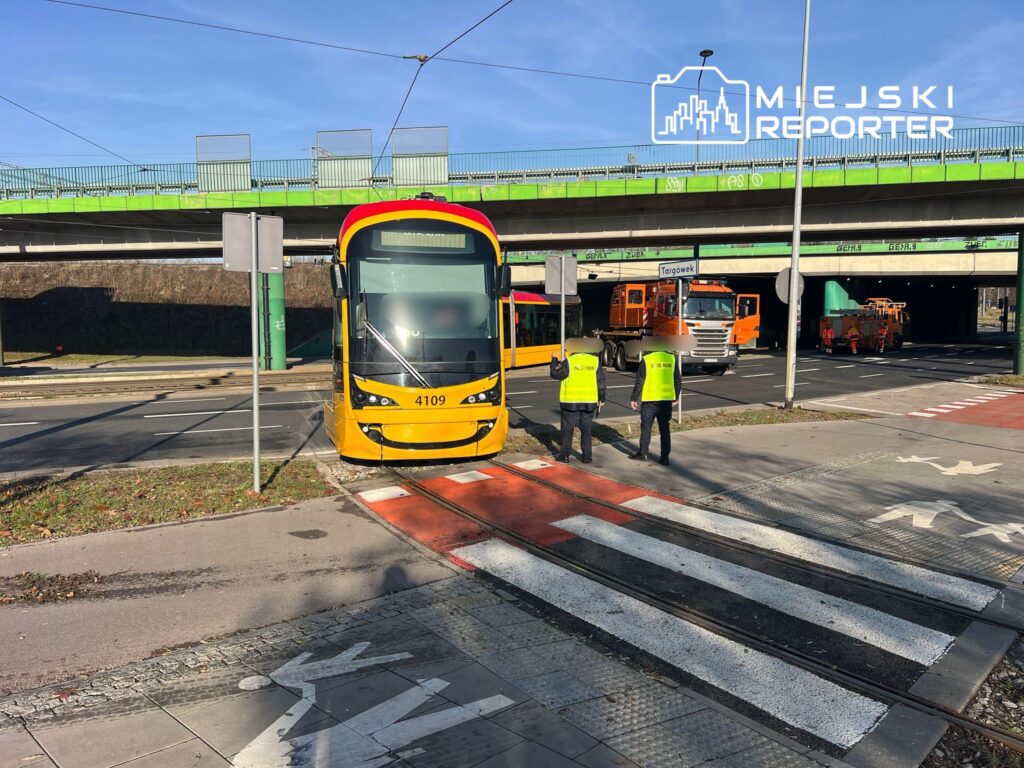 Tramwaj w żółto-czerwonym malowaniu zatrzymany na torowisku, a obok dwóch pracowników w odblaskowych kamizelkach.