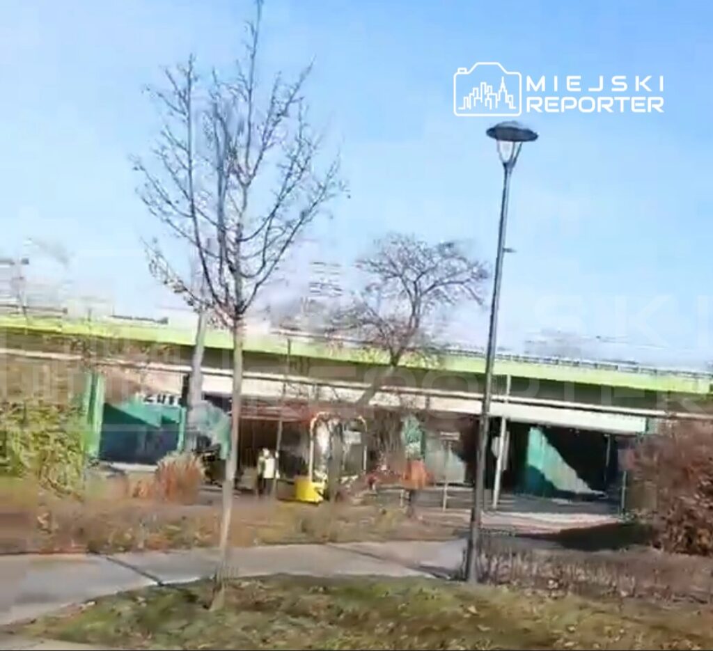 Pojazd poruszający się wzdłuż ścieżki rowerowej obok nowoczesnego wiaduktu, w otoczeniu drzew i lamp ulicznych.