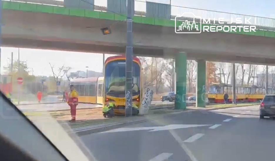 Tramwaj zatrzymany pod wiaduktem, obok pracownicy w odblaskowych kamizelkach, w tle drugi tramwaj na torach.