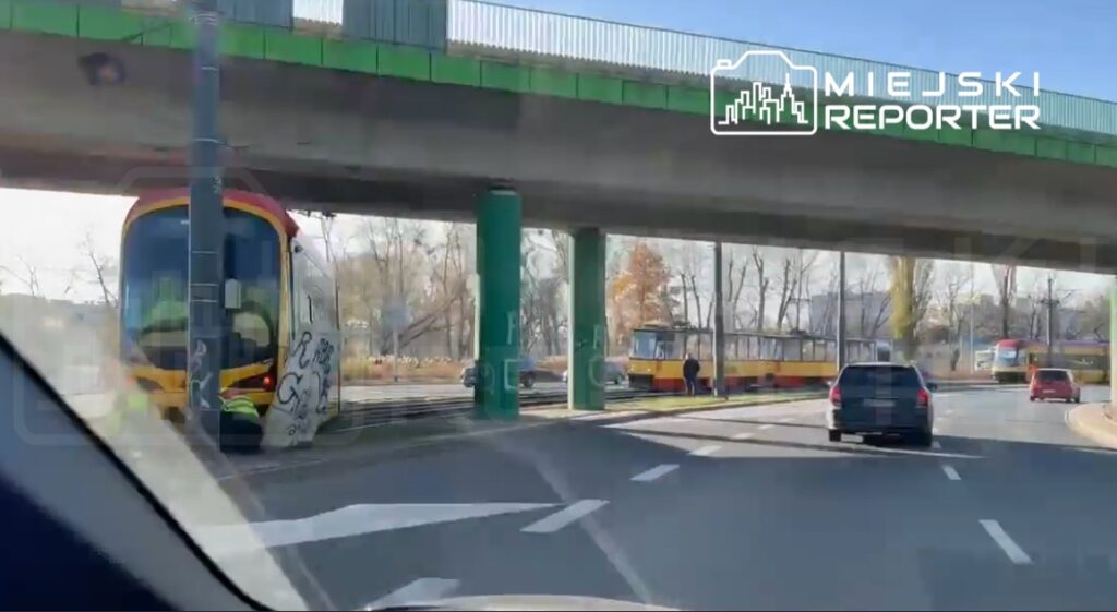 Tramwaje zatrzymane na przystanku obok ruchliwej ulicy, z zielonym wiaduktem w tle.