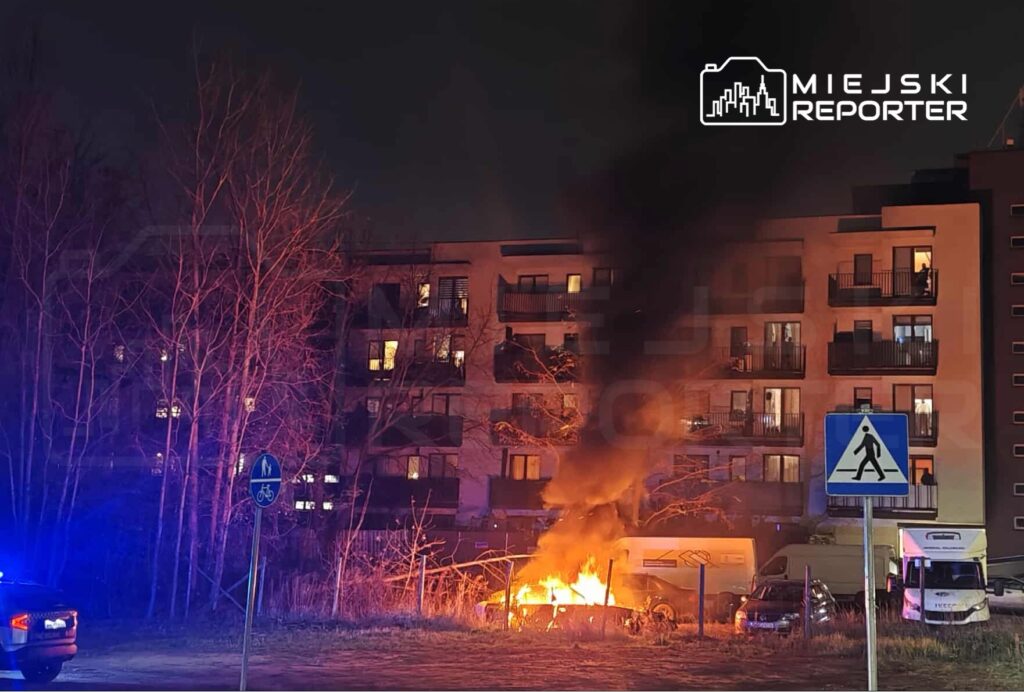 Ogień trawi samochód zaparkowany przy nowoczesnym budynku mieszkalnym, w tle widoczne są zapalone okna.