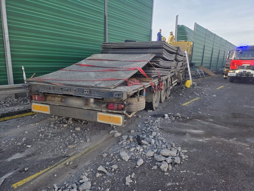 Ciągnik siodłowy z ładunkiem metalowych płyt zatrzymany na drodze, wokół rozrzucone kawałki asfaltu. W tle strażacy interweniują.