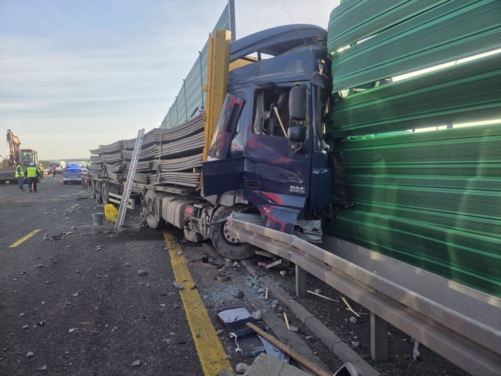 Ciężarówka uderzyła w zieloną barierę dźwiękochłonną na drodze, w otoczeniu zniszczonych elementów jezdni i pojazdów służb.