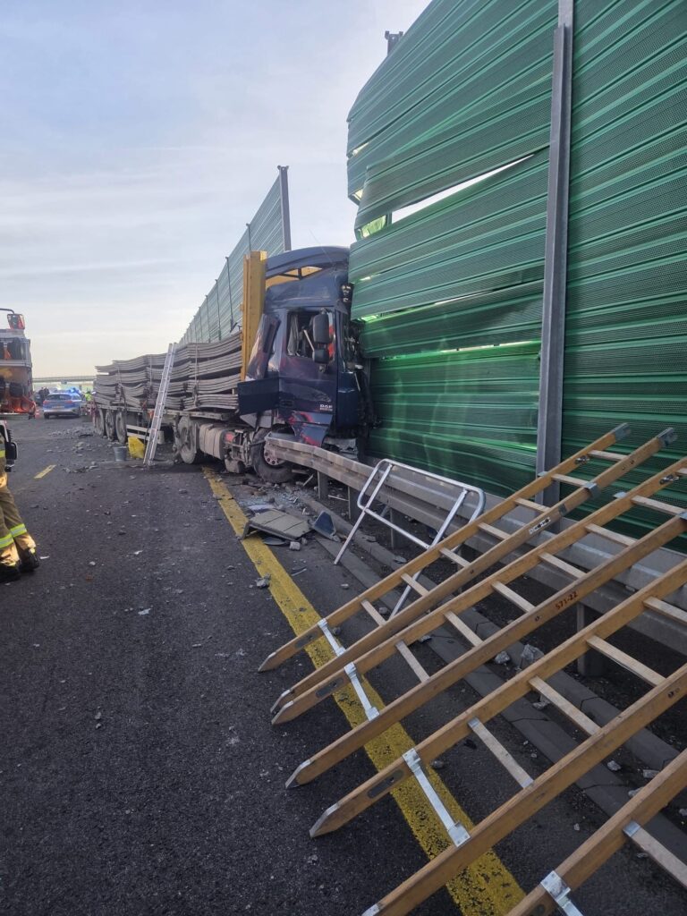 Ciężarówka wjeżdżająca w zieloną barierę dźwiękochłonną, otoczona przez uszkodzone elementy drogi i sprzęt ratunkowy.