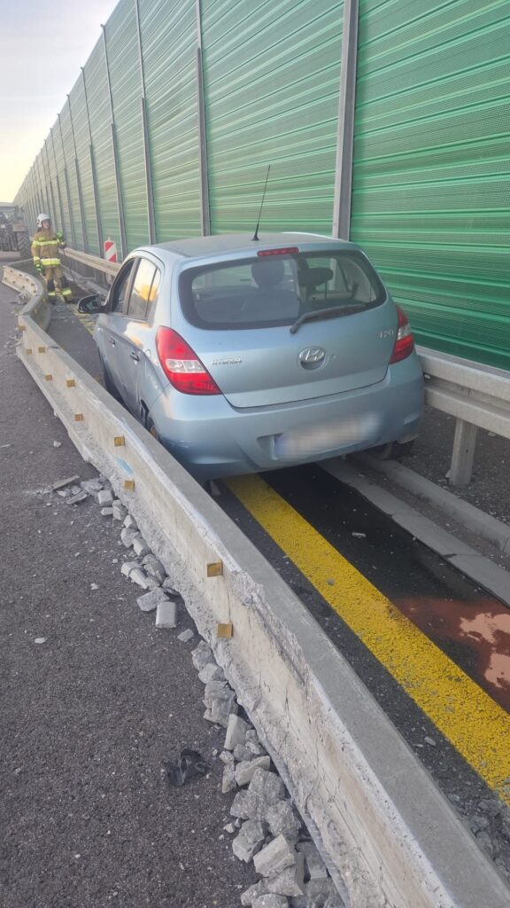 Niebieski samochód osobowy utknął na pasie drogowym obok uszkodzonego betonowego bariery, w tle widać zieloną ścianę akustyczną.