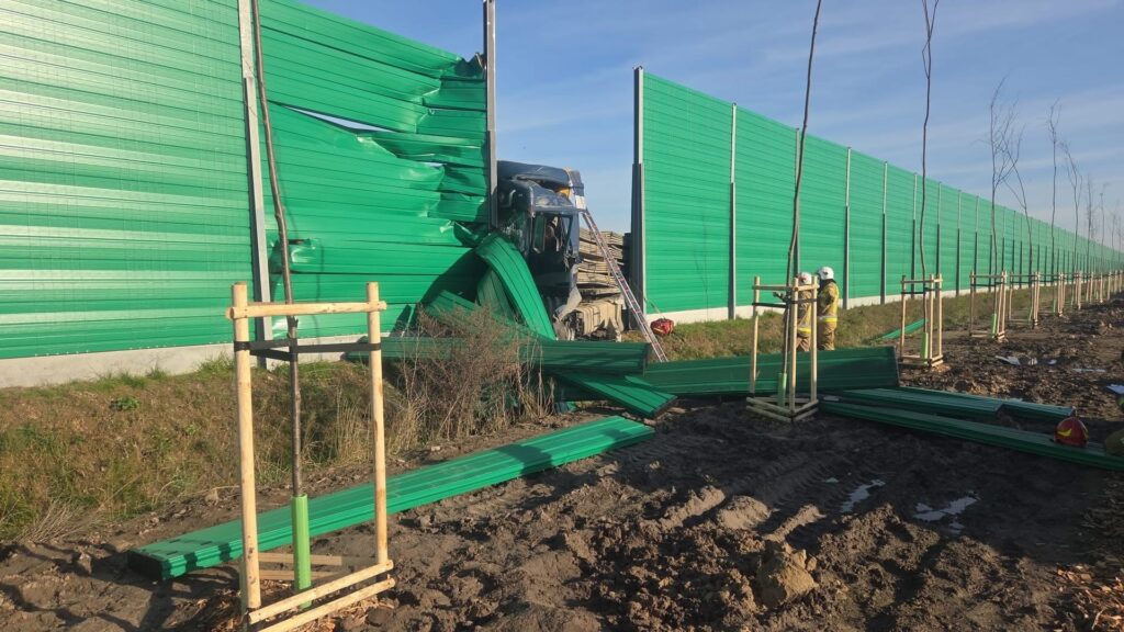 Traktor z uszkodzoną kabiną wjeżdża w zieloną, panelową płot, obok pracują strażacy w kaskach.