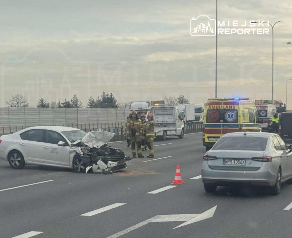 Uszkodzony samochód osobowy z wgniecionym przodem stoi na pasie drogi, a obok pracują strażacy i karetki.
