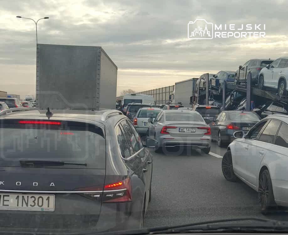 Korki na drodze z wieloma samochodami osobowymi i ciężarówką przewożącą auta w tle. Szare niebo nad ruchliwą trasą.