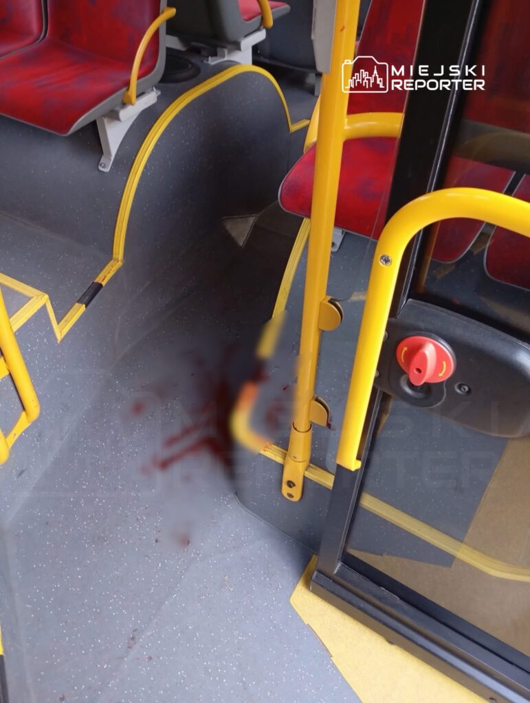 Krew na podłodze wewnątrz autobusu, otoczenie z czerwonymi siedzeniami i żółtymi uchwytami.