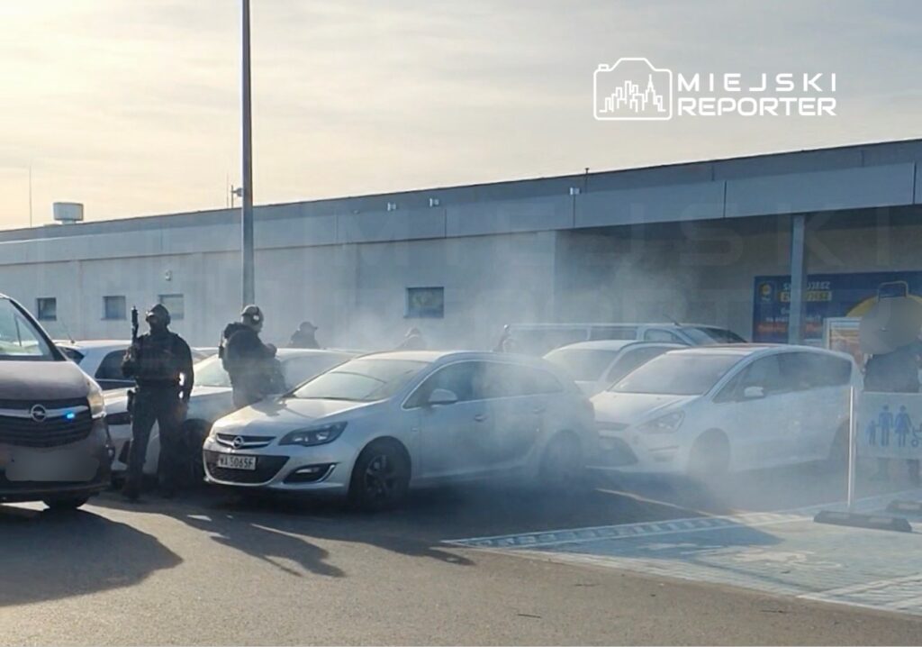 Uzbrojeni funkcjonariusze policji w mundurach interwencyjnych przeszukują parking przed sklepem, otoczonym dymem.