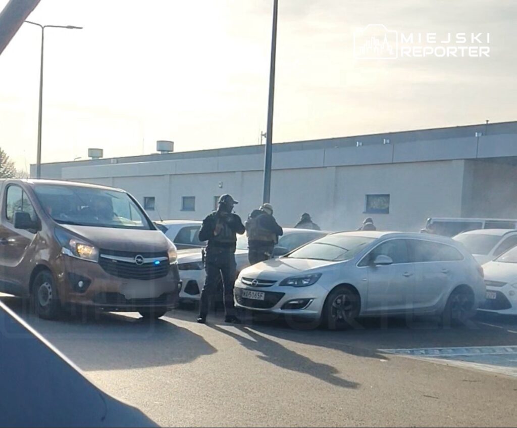 Grupa uzbrojonych funkcjonariuszy policji przeszukuje parking, obok zaparkowanych samochodów.