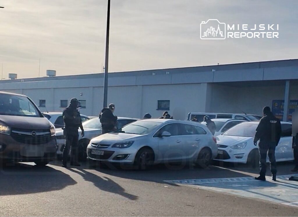 Grupa policjantów w mundurach przeszukuje samochody na parkingu przed budynkiem handlowym.