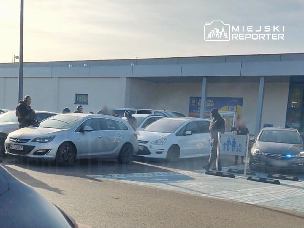 Grupa osób w zewnętrznej części parkingu przed budynkiem handlowym, niektórzy rozmawiają z policjantami.