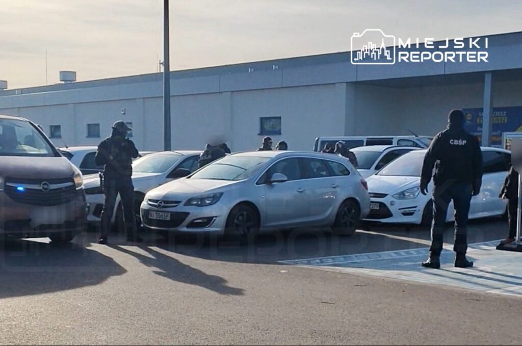 Funkcjonariusze w kamizelkach ochronnych przeszukują zaparkowane samochody na terenie parkingu.