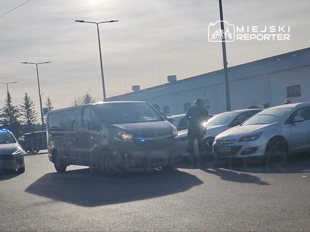 Funkcjonariusz policji rozmawia z mężczyzną obok vanu w parkingu otoczonym zaparkowanymi samochodami.