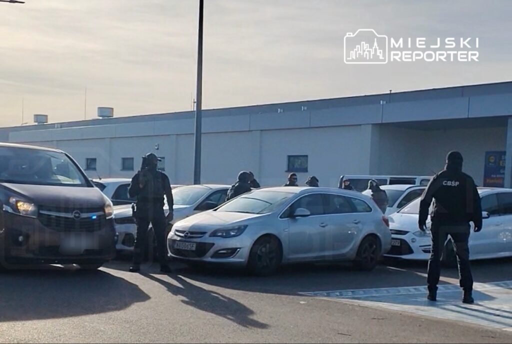 Grupa uzbrojonych funkcjonariuszy w kamizelkach taktycznych przeszukuje parking w pobliżu budynku.