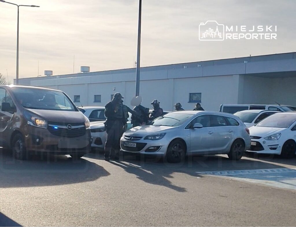 Grupa uzbrojonych policjantów w mundurach rozmawia obok zaparkowanego samochodu w zatłoczonym parkingu.
