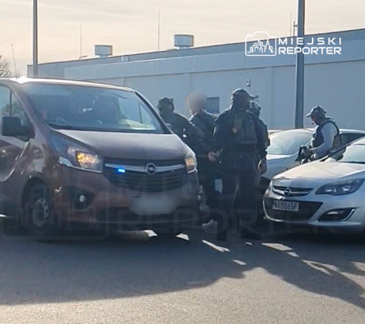 Grupa uzbrojonych funkcjonariuszy otacza van z włączonymi światłami policyjnymi na parkingu.