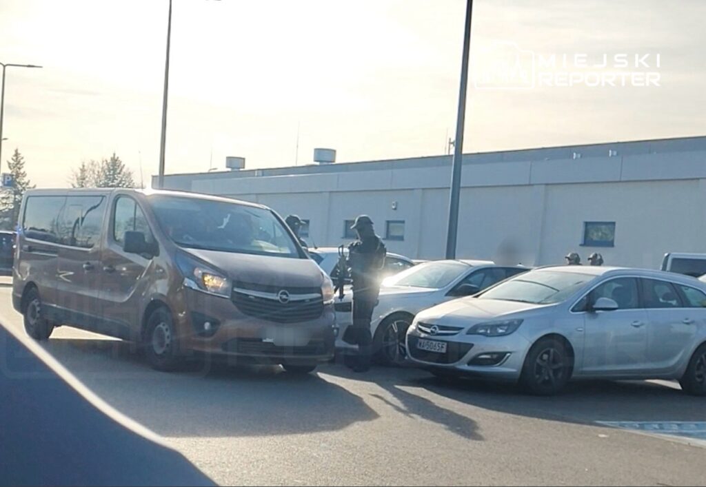 Mundurowy funkcjonariusz z bronią stoi obok furgonetki w strefie parkingowej.