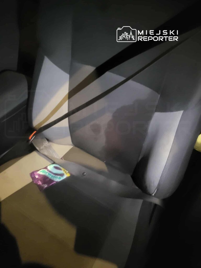 Czarny fotel samochodowy z zapiętym pasem bezpieczeństwa, na siedzeniu leży kolorowa paczka.