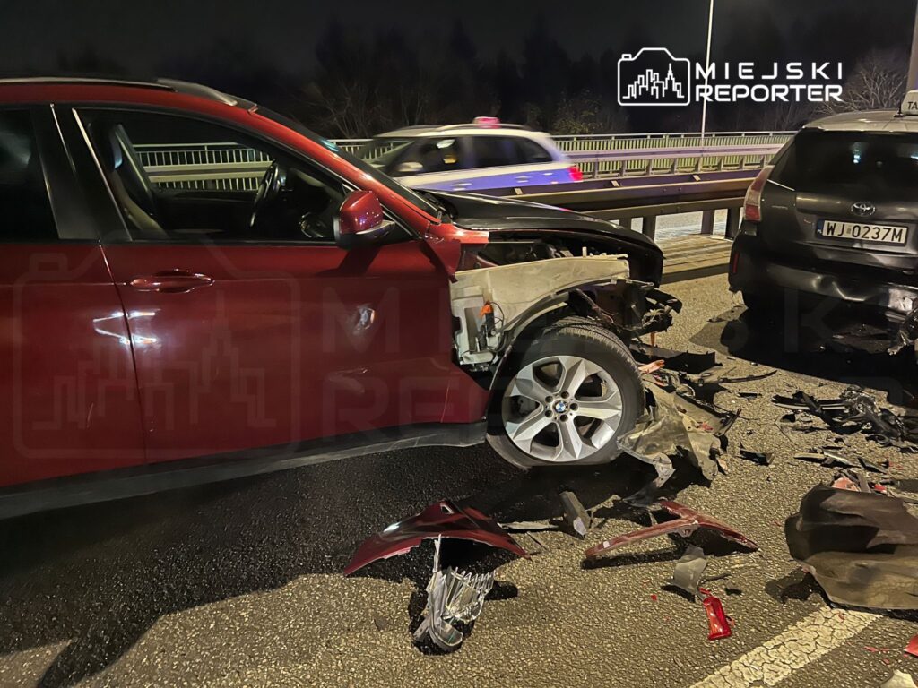 Czerwony samochód osobowy z uszkodzoną przednią częścią stoi na jezdni po kolizji z innym pojazdem w nocy. Wokół leżą fragmenty karoserii.