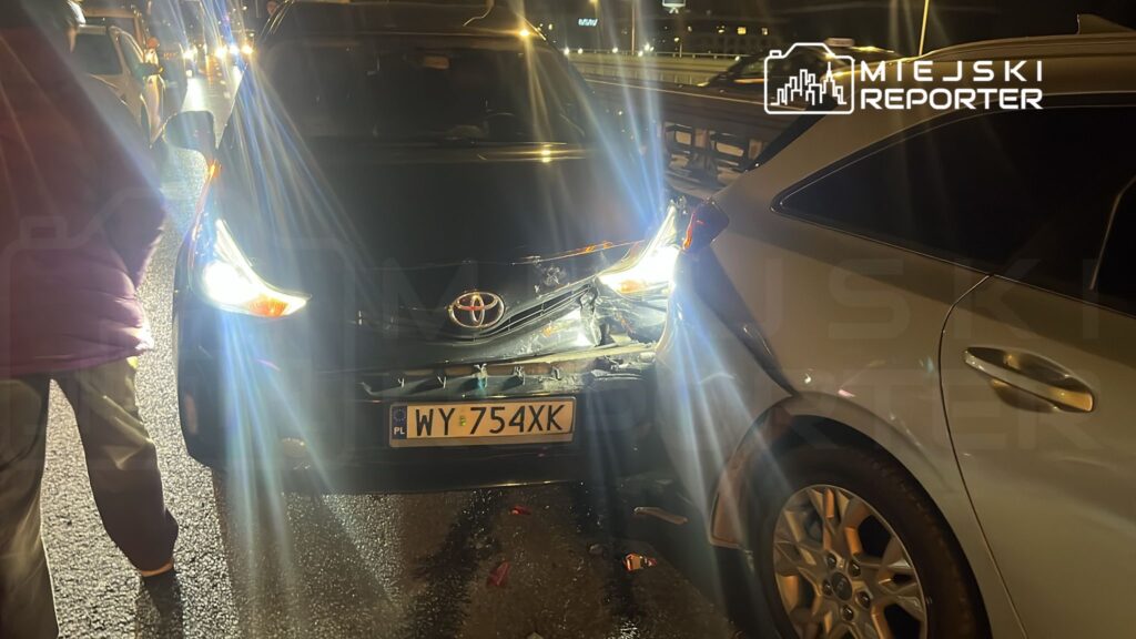 Czarne auto marki Toyota z uszkodzonym tyłem stoi na drodze, obok niego przechodzi osoba w fioletowej kurtce.